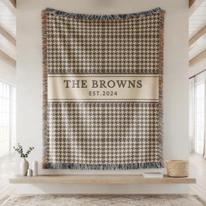 May include: A woven throw blanket featuring a brown and cream houndstooth pattern with a central horizontal band containing the text THE BROWNS and EST.2024. The blanket has fringed edges with multi-colored threads along the vertical sides.