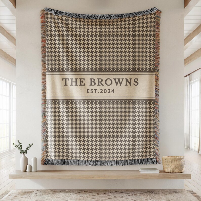 May include: A woven throw blanket featuring a brown and cream houndstooth pattern with a central horizontal band containing the text THE BROWNS and EST.2024. The blanket has fringed edges with multi-colored threads along the vertical sides.