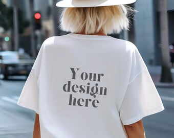 Op de afbeelding: Een wit T-shirt met de tekst "Your design here" gedrukt op de achterkant. Het shirt wordt gedragen door een persoon met blond haar en een witte hoed.