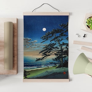 Puede incluir: Una impresi&oacute;n de una escena costera iluminada por la luna con un gran pino, olas y una playa de arena. La obra de arte est&aacute; montada en barras de madera y cuelga de un cord&oacute;n negro. Tambi&eacute;n se ven un rollo enrollado y una peque&ntilde;a planta en maceta.
