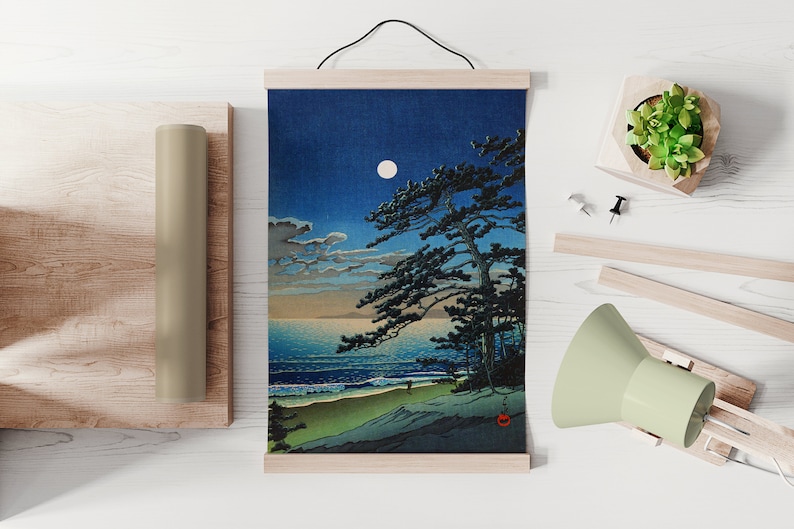 Puede incluir: Una impresi&oacute;n de una escena costera iluminada por la luna con un gran pino, olas y una playa de arena. La obra de arte est&aacute; montada en barras de madera y cuelga de un cord&oacute;n negro. Tambi&eacute;n se ven un rollo enrollado y una peque&ntilde;a planta en maceta.
