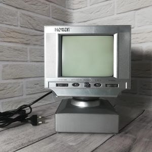 May include: A silver portable television set with a small screen and the brand name PRESIDENT printed above the display. The device features a square base, a black power cord with a plug, and manual control dials for volume and tuning.