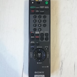 May include: Black remote control for a Sony video player. The remote has buttons for play, pause, stop, rewind, fast forward, and other functions. It also has a number pad and buttons for volume, program, and input select.