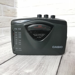 Pode incluir: Um toca-fitas de rádio estéreo AM/FM Casio cinza escuro. O rádio possui um dial de sintonia com frequências FM e AM, e o número do modelo AS-250R é visível. A parte superior possui três botões de controle.