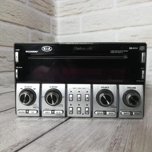 Może przedstawiać: Czarny i srebrny samochodowy zestaw stereo z logo Kia i Clarion. Zestaw stereo ma kilka przycisków i pokręteł do funkcji takich jak wyszukiwanie, głośność i źródło. Widoczny jest numer modelu WX2468RMP.