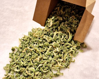 May include: A brown cardboard box, partially open, spilling out a pile of dried, green, and yellow-green vegetable pieces. The pieces are small and have a textured, ridged appearance. The box is on a white, textured surface.