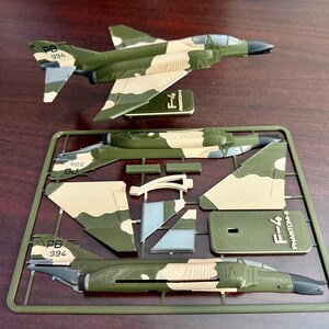 以下が含まれることがあります： オリーブグリーンとタンの迷彩柄のF-4ファントムIIモデルキット。キットには、未組立のパーツ、完成したモデル、ベースが含まれています。モデルの尾翼には「994」の数字がプリントされています。キットは濃い茶色の表面にあります。