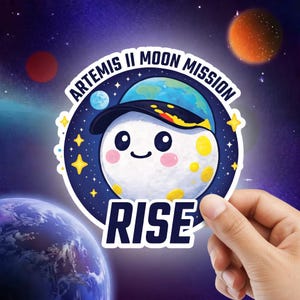 Puede incluir: Una pegatina con una luna de dibujos animados que lleva una gorra, con el texto "ARTEMIS II MOON MISSION" y "RISE". La luna tiene una cara sonriente y está rodeada de estrellas y planetas sobre un fondo espacial.
