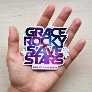 Puede incluir: Una pegatina blanca con las palabras "GRACE ROCKY SAVE STARS" en una fuente con temática espacial, con un diseño de galaxia púrpura y azul. El texto "PROJECT HAIL MARY" está en la parte inferior. La pegatina se sostiene en una mano.