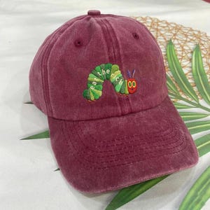 Könnte beinhalten: Burgunderfarbene Baseballkappe mit gebogenem Schirm. Die Kappe hat ein gesticktes Design einer grünen und gelben Raupe mit dem Text "the very rich". Die Kappe ist aus einem weichen, strukturierten Stoff gefertigt.
