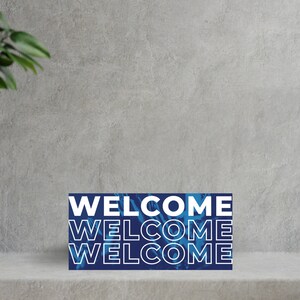 K&ouml;nnte beinhalten: Ein blaues und wei&szlig;es Willkommensschild mit dem Wort "WELCOME", das dreimal wiederholt wird. Das Schild befindet sich auf einer grauen Oberfl&auml;che mit gr&uuml;nen Pflanzen auf beiden Seiten.