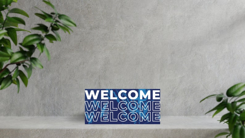 K&ouml;nnte beinhalten: Ein blaues und wei&szlig;es Willkommensschild mit dem Wort "WELCOME", das dreimal wiederholt wird. Das Schild befindet sich auf einer grauen Oberfl&auml;che mit gr&uuml;nen Pflanzen auf beiden Seiten.