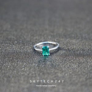 Peut inclure: Une bague en argent ornée d'une pierre verte rectangulaire taille émeraude. La bague est posée sur une surface texturée gris foncé. Le texte "SKYTECH 247 Handcrafted Jewellery" est en bas.