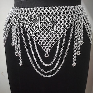 Puede incluir: Un cinturón de cota de malla plateada con un patrón geométrico y cadenas colgantes. El cinturón presenta el texto "Chained Body Jewellery". Se muestra en un maniquí negro.