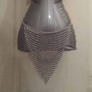 Peut inclure: Un vêtement en cotte de mailles argentée, probablement une pièce d'armure, est suspendu à un mannequin. La cotte de mailles est constituée de petits anneaux métalliques imbriqués, formant une forme triangulaire. Le mannequin est gris.