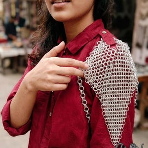 Puede incluir: Una hombrera de cota de malla plateada con un patrón triangular de anillos metálicos entrelazados. El accesorio está sujeto al hombro de una camisa abotonada de color rojo oscuro mediante un pequeño botón metálico y una cadena vertical.
