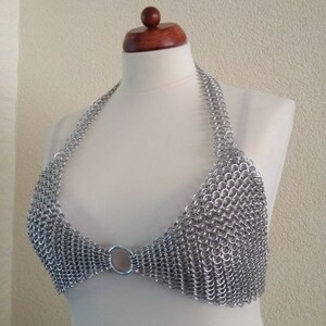 Puede incluir: Top halter de cota de malla plateada exhibido en un maniquí. La parte superior está hecha de pequeños anillos de metal entrelazados, formando una tela texturizada y metálica. Las correas halter y la parte delantera presentan un diseño circular.