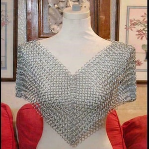 Puede incluir: Una parte superior de cota de malla plateada exhibida en un maniquí. La cota de malla está hecha de pequeños anillos de metal entrelazados, formando un diseño de cuello en V. La parte superior sin mangas parece ser una pieza para los hombros o una capa.