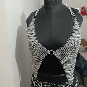 May include: A silver chainmail top displayed on a black mannequin. The halter-style top is constructed of small, interlocking metal rings, creating a mesh-like appearance. The design features a plunging neckline and a backless style, showcasing a unique, handcrafted aesthetic.
