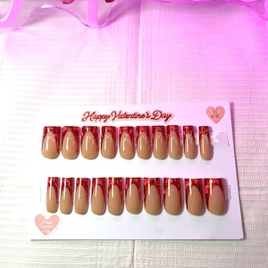 May include: A set of artificial nails in a neutral beige color with a red glitter design. The nails are arranged on a white card with the text "Happy Valentine's Day" and a heart-shaped sticker that says "Be Mine".