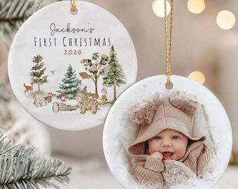 May include: Two round white ceramic ornaments with gold hanging strings. One features a watercolor forest scene with a bear, deer, and the text Jackson's FIRST CHRISTMAS 2026. The other displays a photograph of a baby wearing a tan hooded sweater.