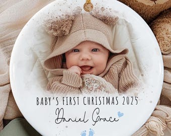 May include: A round white ceramic ornament featuring a photograph of a baby wearing a tan hooded sweater. The text reads "BABY'S FIRST CHRISTMAS 2025" and "Daniel Grace" in black script, with two small blue footprints and a blue heart icon below.