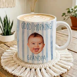 May include: White ceramic mug with a blue striped design, the words "GOOD MORNING GRANDPA", and a baby's face. The mug sits on a cream-colored macrame coaster on a wooden table. The mug is filled with a latte.