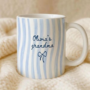 May include: A white ceramic mug with blue vertical stripes. The mug has a white handle and features the text "Olivia's grandma" in a script font, with a bow illustration. The mug is set against a soft, off-white knitted background.