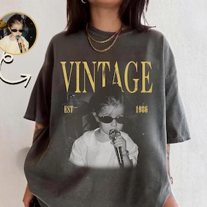 May include: A charcoal gray oversized t-shirt featuring a graphic print of a child wearing dark sunglasses and holding a microphone. The text VINTAGE EST 1986 is printed in yellow above and below the black and white photographic image.