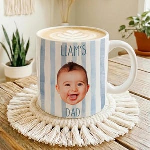 May include: White ceramic mug with blue vertical stripes, featuring a baby's photo and the text "LIAM'S DAD." The mug is filled with a latte and rests on a cream-colored macrame coaster. The mug is on a wooden table.