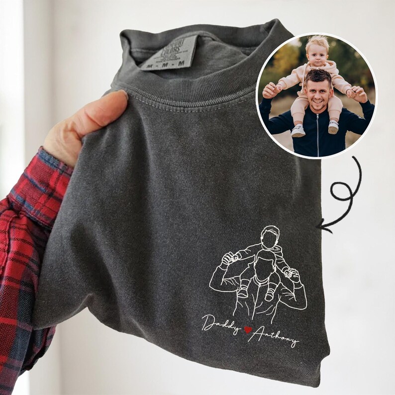 May include: Dark gray t-shirt with a white line drawing of a person carrying a child on their shoulders. The text "Daddy Anthony" is below the drawing, with a red heart. A small photo of a person and child is in the upper right corner.