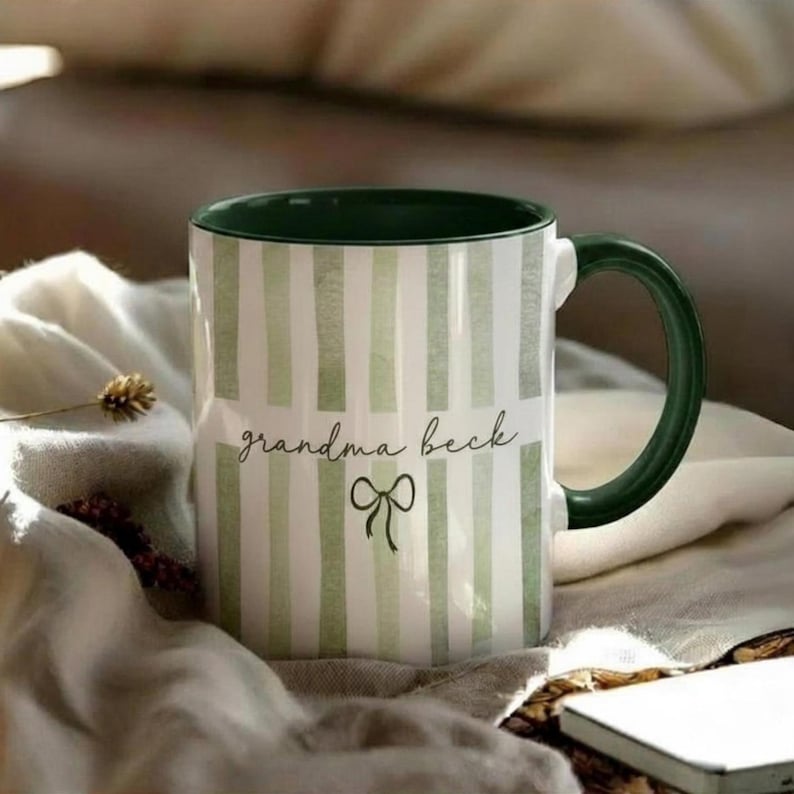 May include: A white ceramic mug with green vertical stripes and a green handle and rim. The mug has the text "grandma beck" in a script font with a bow graphic. The mug is on a white cloth.