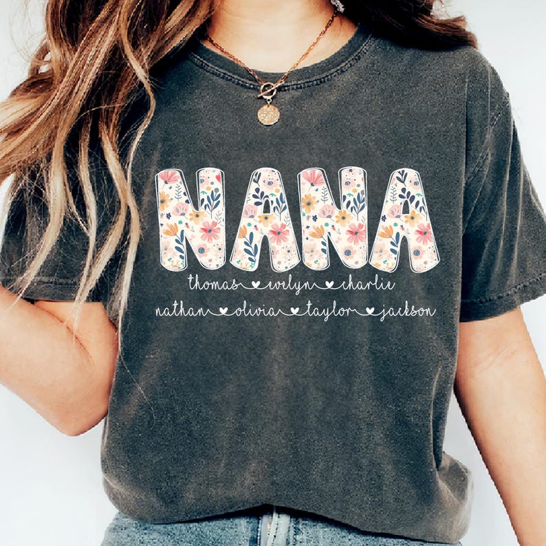 May include: Dark gray t-shirt with the word "NANA" in large, floral-patterned letters. Below the word are names: thomas, evelyn, charlie, nathan, olivia, taylor, and jackson. The shirt has a crew neck.