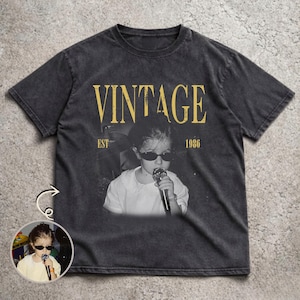 May include: A charcoal gray short-sleeve t-shirt featuring a faded graphic print of a child wearing sunglasses and holding a microphone. The text VINTAGE is printed in large yellow letters above the image, with EST 1986 printed below the text.