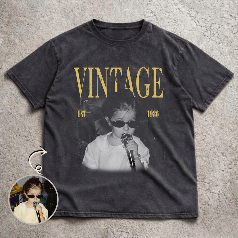 May include: A charcoal gray short-sleeve t-shirt featuring a faded graphic print of a child wearing sunglasses and holding a microphone. The text VINTAGE is printed in large yellow letters above the image, with EST 1986 printed below the text.
