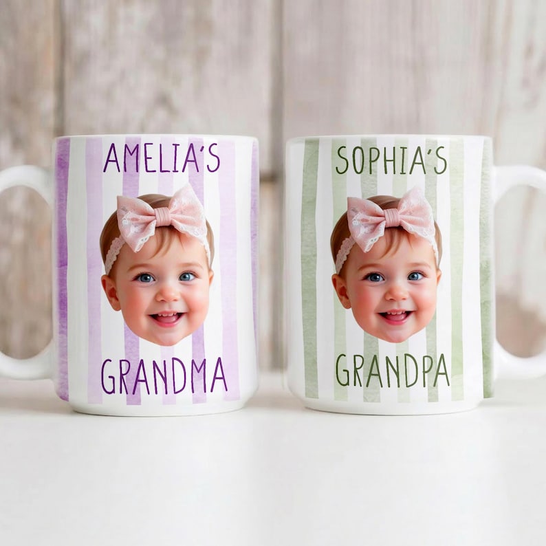 May include: Two white ceramic mugs with handles. One mug has purple stripes and the text "AMELIA'S GRANDMA" with a baby's photo. The other mug has green stripes and the text "SOPHIA'S GRANDPA" with a baby's photo.