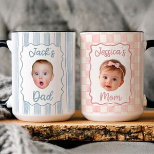 May include: Two ceramic mugs with black handles. One mug features a baby's photo with the text "Jack's Dad" on a blue striped background. The other mug has a baby's photo with the text "Jessica's Mom" on a pink checkered background.
