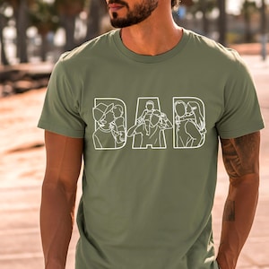 Peut inclure: T-shirt vert olive avec le mot "DAD" en blanc, avec des illustrations au trait &agrave; l'int&eacute;rieur de chaque lettre. Les illustrations repr&eacute;sentent un parent avec un enfant, cr&eacute;ant un design sur le th&egrave;me de la famille. Le t-shirt est &agrave; manches courtes.
