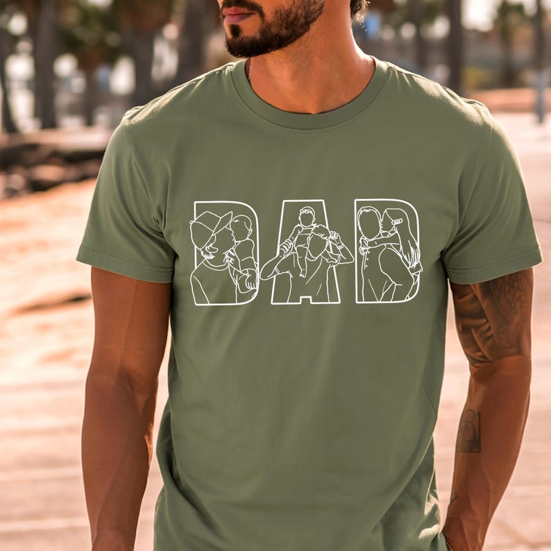 Peut inclure: T-shirt vert olive avec le mot "DAD" en blanc, avec des illustrations au trait &agrave; l'int&eacute;rieur de chaque lettre. Les illustrations repr&eacute;sentent un parent avec un enfant, cr&eacute;ant un design sur le th&egrave;me de la famille. Le t-shirt est &agrave; manches courtes.