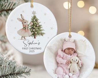 May include: Two round white ceramic ornaments with gold hanging strings. One features a watercolor illustration of a rabbit in a pink tutu decorating a Christmas tree with the text Sophia's FIRST CHRISTMAS 2025. The other shows a photo of a baby.