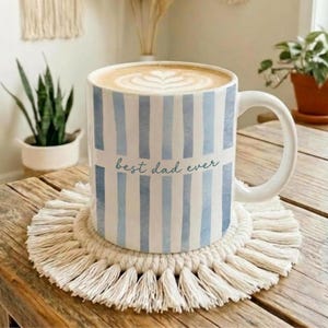 May include: A white ceramic mug with blue vertical stripes and the text "best dad ever." The mug sits on a cream-colored macrame coaster. The mug is filled with a latte with a heart design.