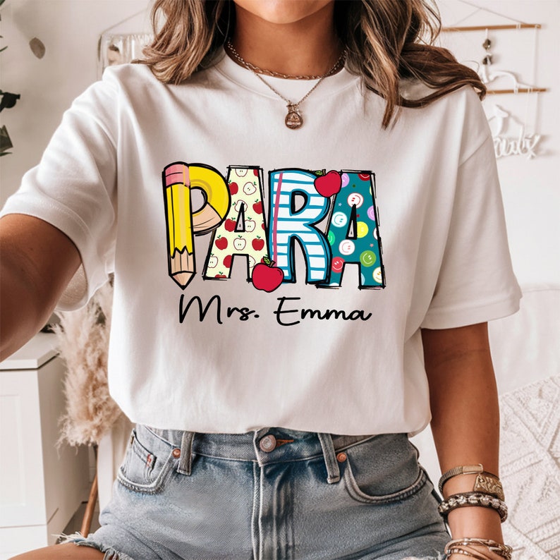 以下が含まれることがあります： カラフルなグラフィックがプリントされた白いTシャツ。グラフィックには「PARA」の文字と、鉛筆、リンゴ、その他の文房具が描かれています。「Mrs. Emma」という文字がグラフィックの下にあります。