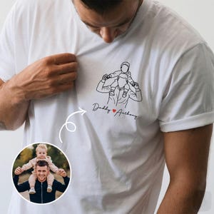 May include: White t-shirt featuring a line drawing of a person carrying a child on their shoulders. The text "Daddy ❤️ Anthony" is below the drawing. A photo of a person with a child on their shoulders is in a circle.