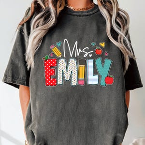 May include: A dark gray t-shirt with the text "Mrs. Emily" in colorful letters. The letters are decorated with school-themed designs, including pencils, apples, and polka dots.