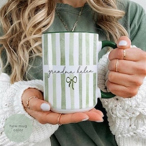 May include: A white ceramic mug with green vertical stripes and a green handle. The mug features the text "grandma helen" in cursive with a green bow graphic. The words "new mug color" are in a circle in the lower left.