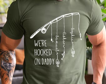 May include: Olive green t-shirt with a white graphic of a fishing rod and the text "WE'RE HOOKED ON DADDY". The names Samuel, Asher, and Sophia are written on the fishing lines, with a fish at the end of each line. A unique gift for Father's Day.