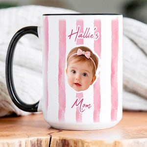 May include: A white ceramic mug with a black handle and rim, featuring vertical pink watercolor stripes. The front displays a photograph of a baby wearing a pink bow headband, with the text Hallie's Mom printed in a pink script font above and below.