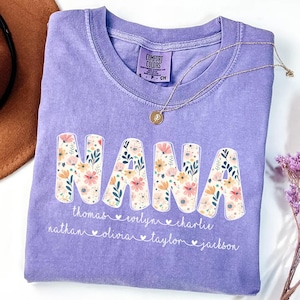May include: A lilac Comfort Colors t-shirt with the word "NANA" in large, floral-filled letters. Below the word are names, including Thomas, Evelyn, and Charlie. A gold necklace with a pendant rests on the shirt.