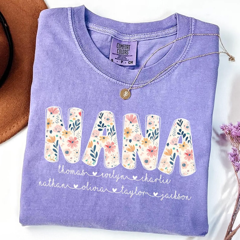 May include: A lilac Comfort Colors t-shirt with the word "NANA" in large, floral-filled letters. Below the word are names, including Thomas, Evelyn, and Charlie. A gold necklace with a pendant rests on the shirt.