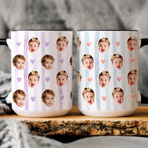 May include: Two ceramic mugs with black handles and rims. One mug features a lavender striped pattern with baby faces and purple hearts. The other mug has a blue striped pattern with baby faces and pink hearts. Both mugs are on a wooden surface.